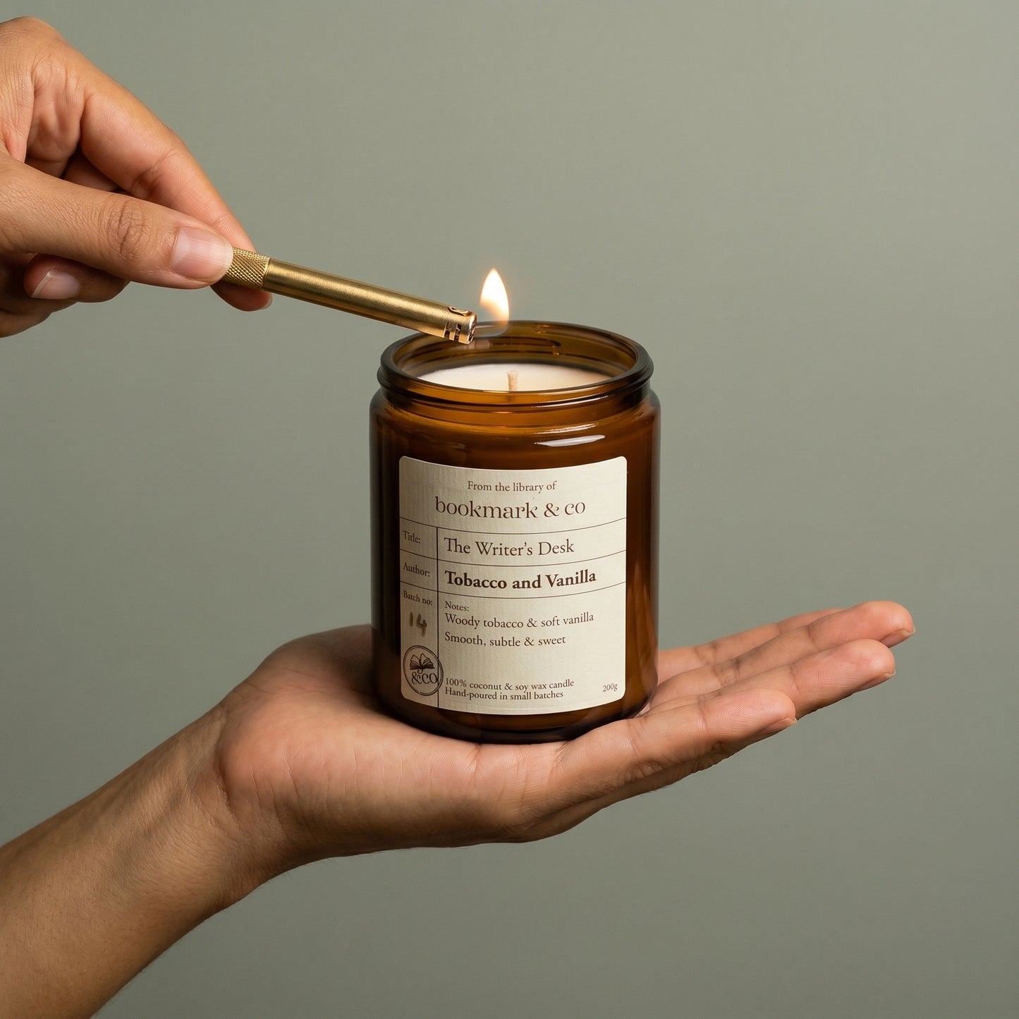 Hand lighting a candle labeled 'The Writer's Desk' by Bookmark & Co on a neutral background