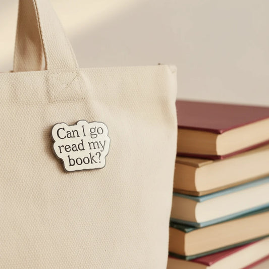 Enamel pin with text 'Can I go read my book?' on a pink and white background