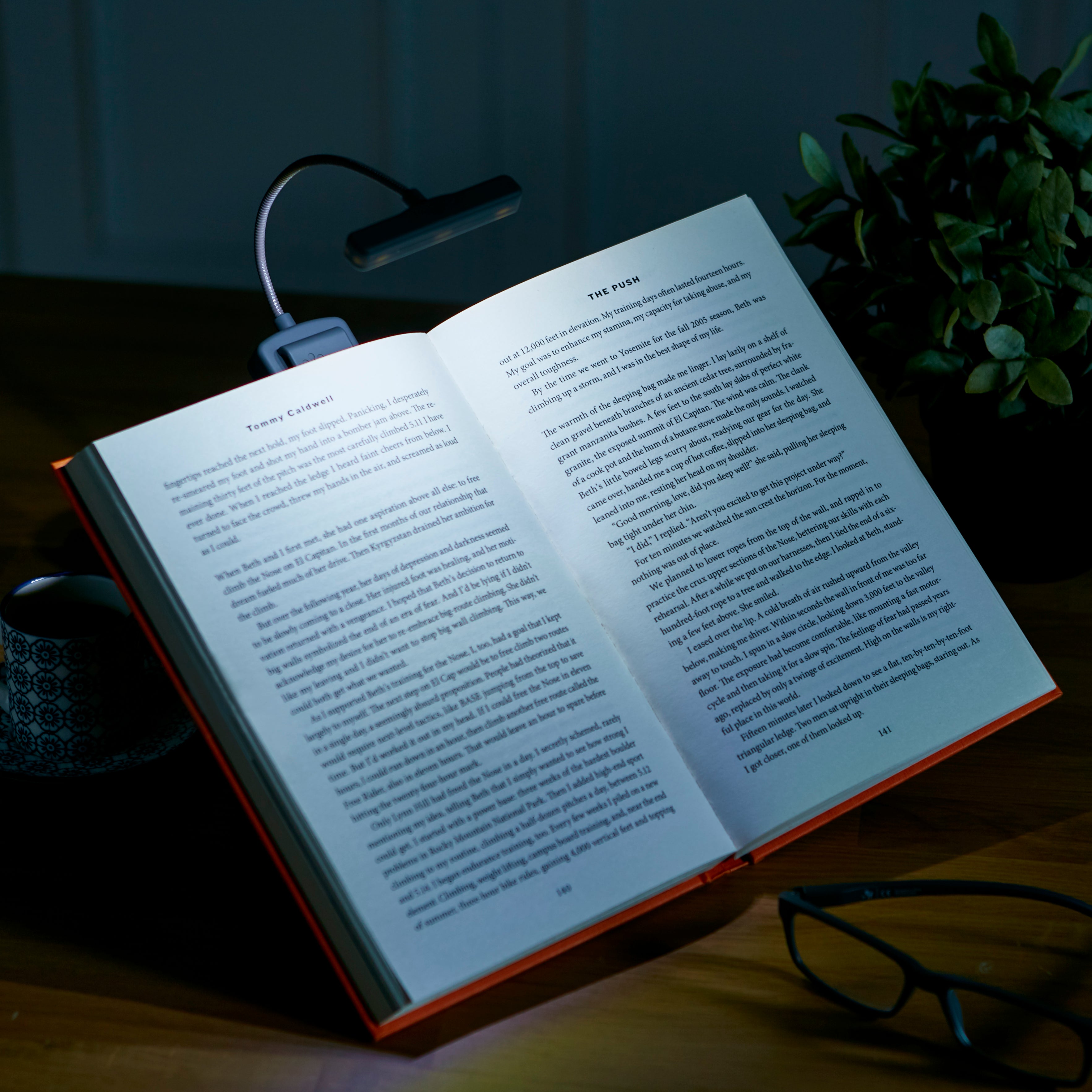 Open book on a table with a lamp illuminating it, next to a mug and glasses.