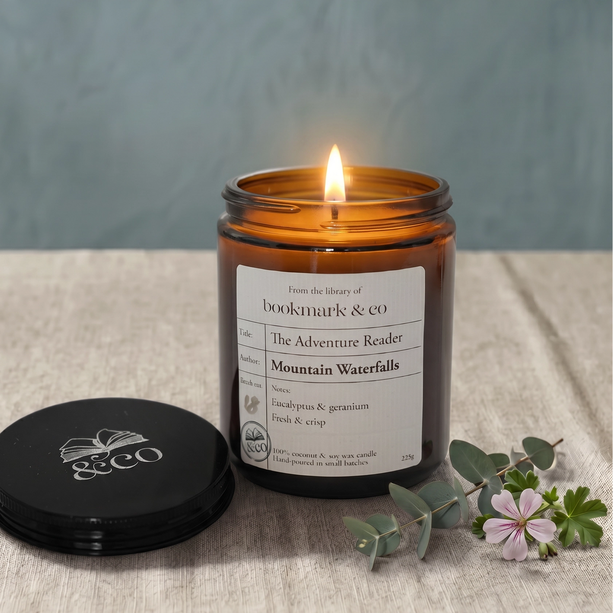 Candle in an amber jar with a label on a textured surface, surrounded by greenery and flowers.