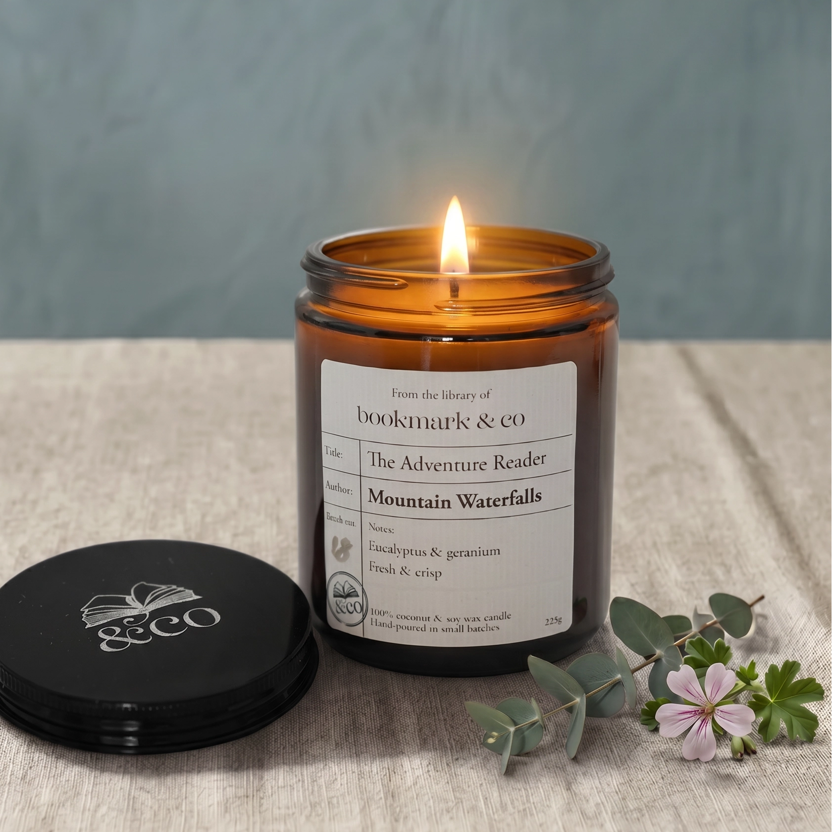 Candle in an amber jar with a label on a textured surface, surrounded by greenery and flowers.