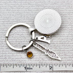 Silver keychain with round textured disc and small knife-shaped charm on a paper background with text.