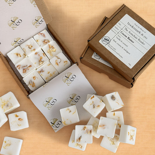 Open and closed kraft gift boxes containing white square wax melts with dried botanical inclusions, arranged on a peach fabric background. The label reads 'The Artisan Reader' and 'Cocoa Butter'.