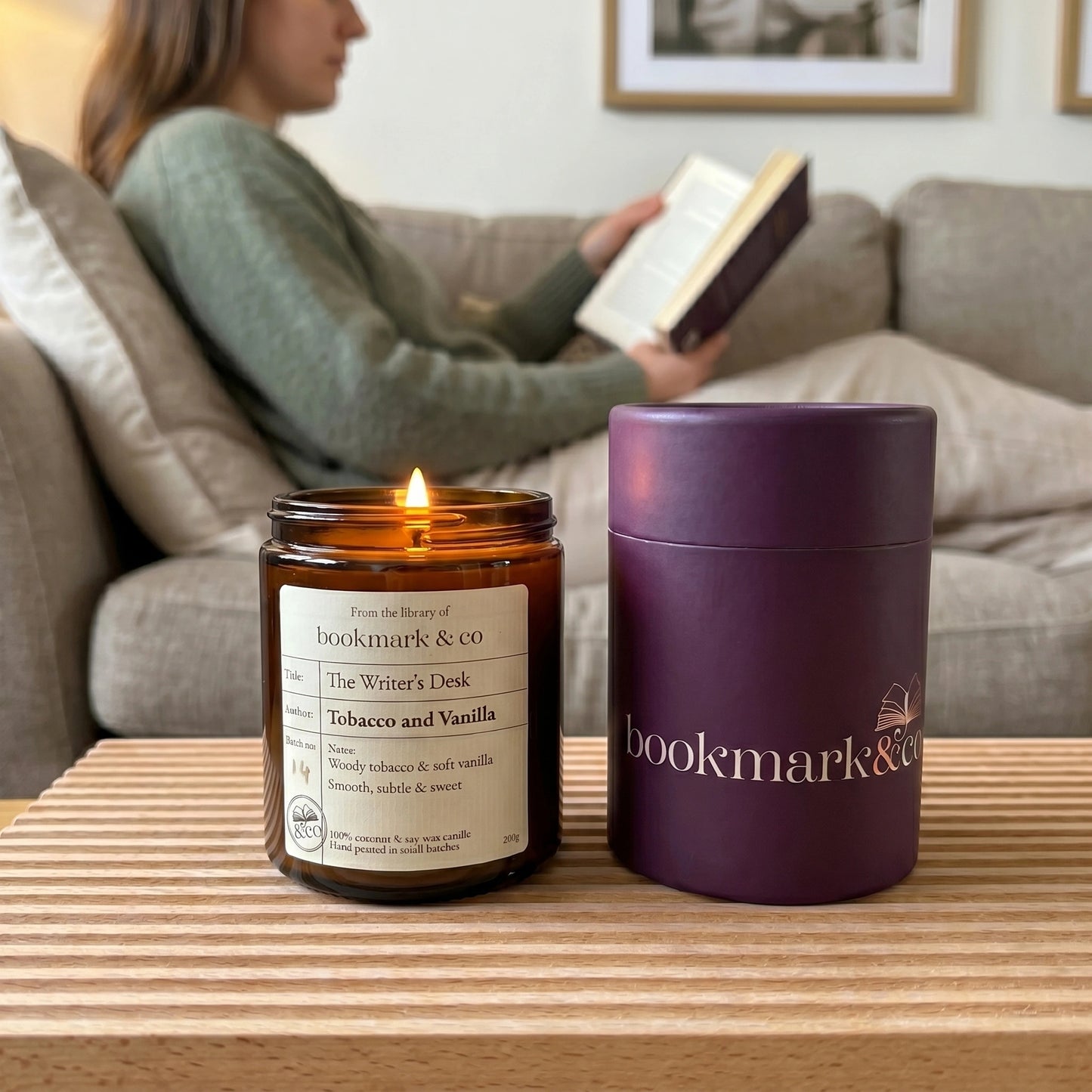 Candle and purple container with 'bookmark & co' branding on a table, person reading book in background