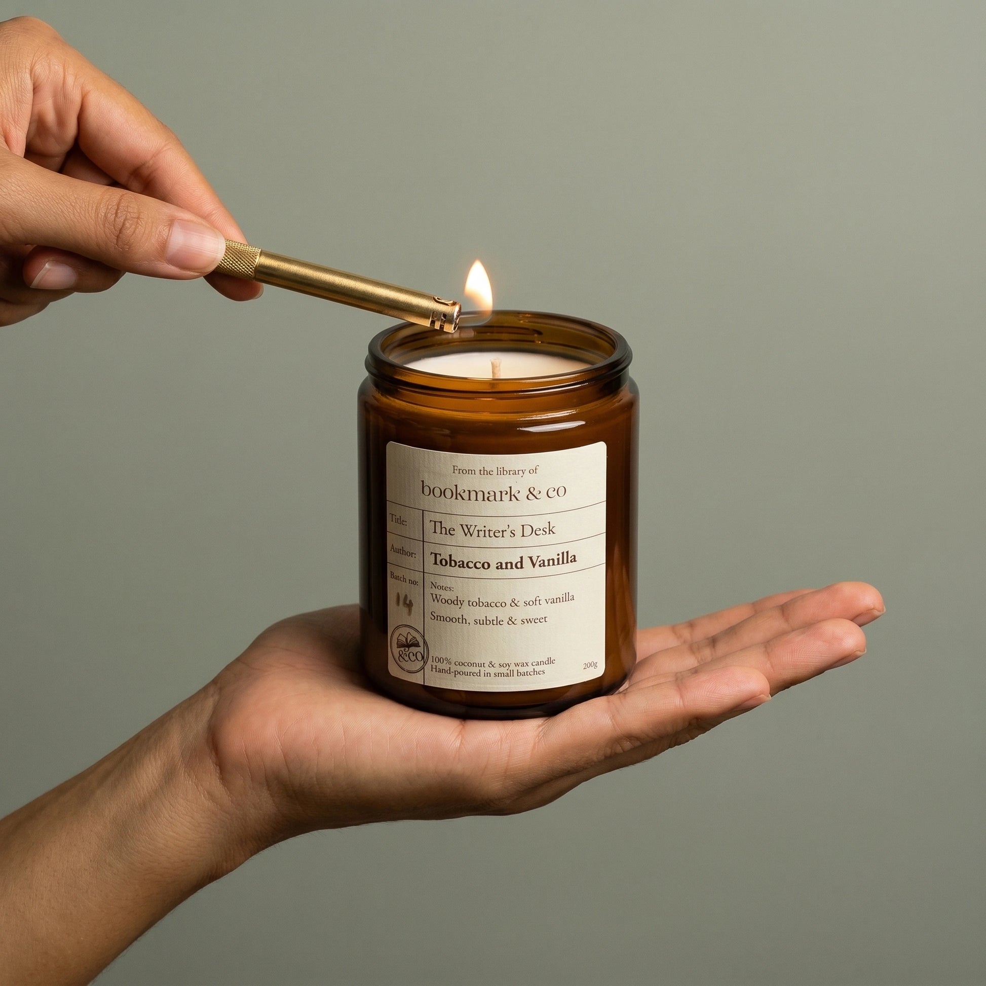 Hand lighting a candle labeled 'The Writer's Desk' by Bookmark & Co on a neutral background