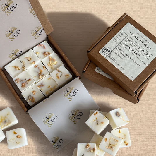 White square wax melts with dried botanical pieces, shown in an open white box alongside a brown kraft box labeled 'Cinnamon Buns' and branded 'bookworms & co'.