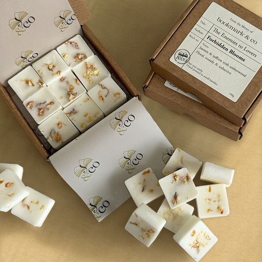 White wax melts with dried flower petals in a brown cardboard box, with several wax melts spilled out on a beige surface. A second box lid is shown with a printed label reading 'The Enemies to Lovers' 'Forbidden Blooms' from 'Bookmark & Co'.