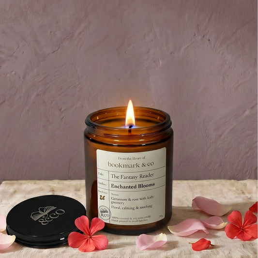 Candle in an amber jar with a label, surrounded by pink flowers on a textured surface.