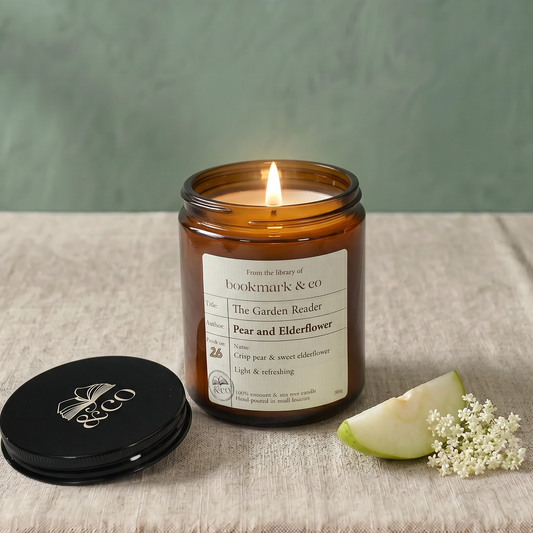 Candle in an amber jar with a label, next to a green apple and white flowers on a textured surface.