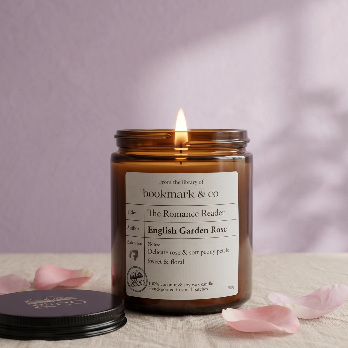 Lit candle in a jar with a label on a textured surface with pink petals, against a soft purple background.