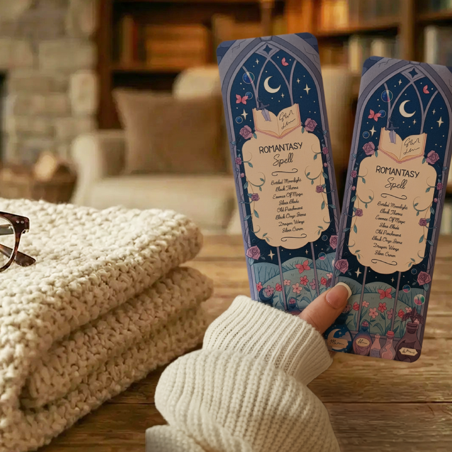 Two  'Romantasy Spell' bookmarks with decorative designs being held in a cosy scene