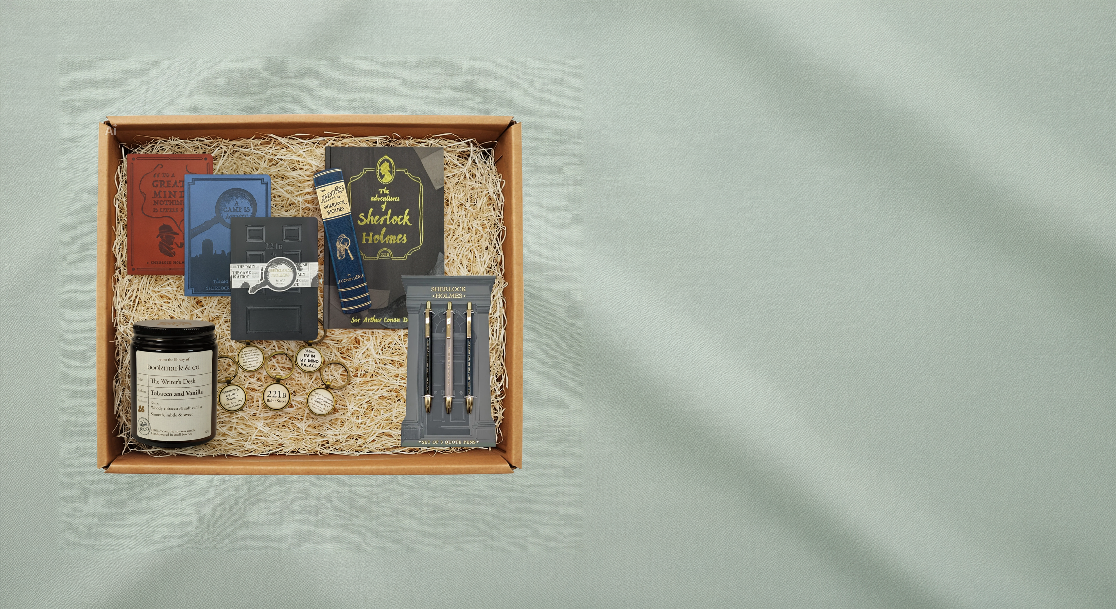 A curated gift box set in a wooden tray filled with shredded paper, containing a candle, small circular items, a black pouch, a blue card, and a gray pen set, photographed from above on a pale green background.