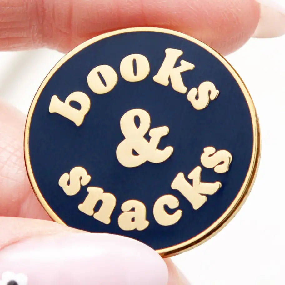 Round enamel pin with 'books & snacks' text held between fingers