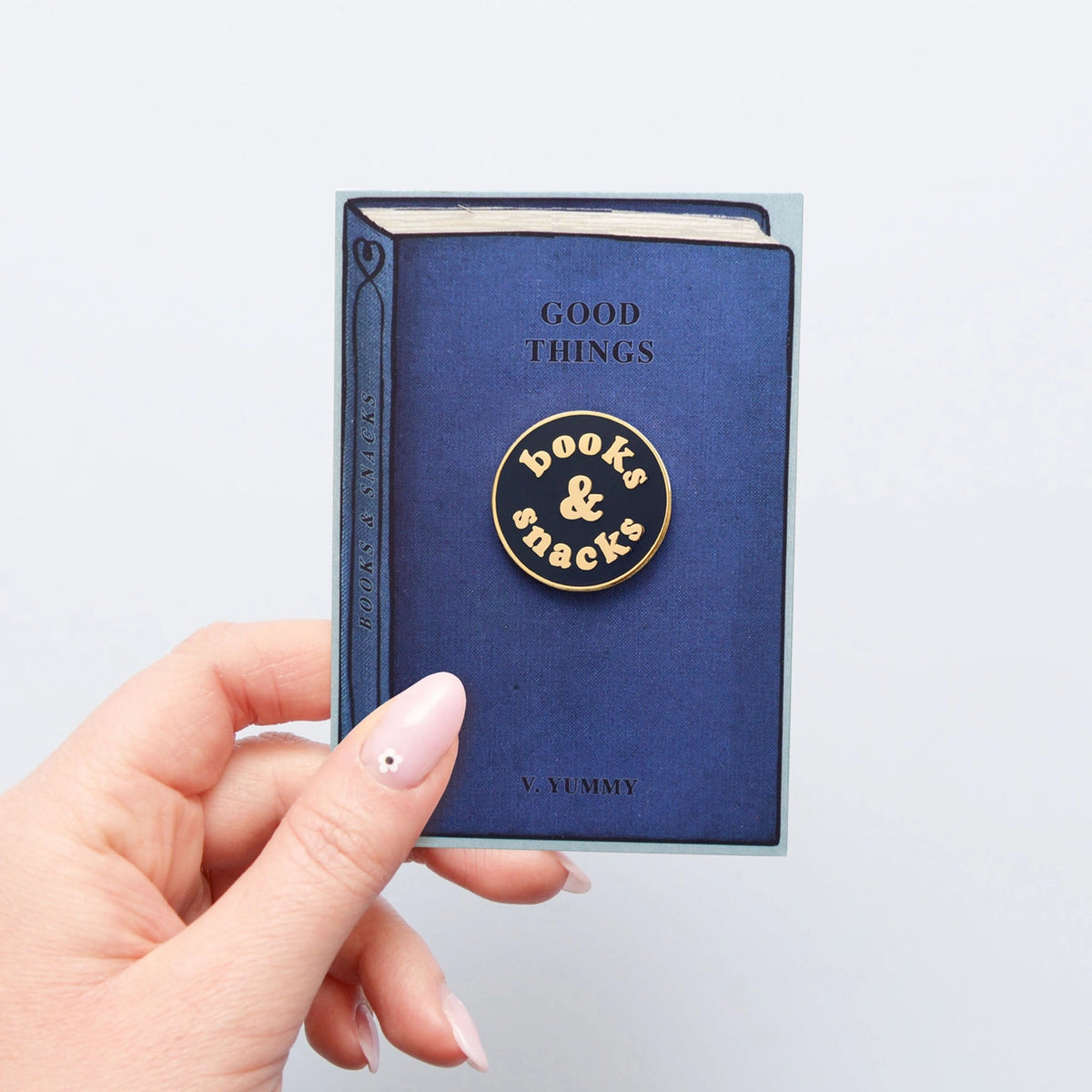 Blue book with 'Good Things' text and 'booke & snacks' logo held by a hand on a light gray background