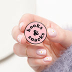 Hand holding a round pink badge with 'books & snacks' text on a light background