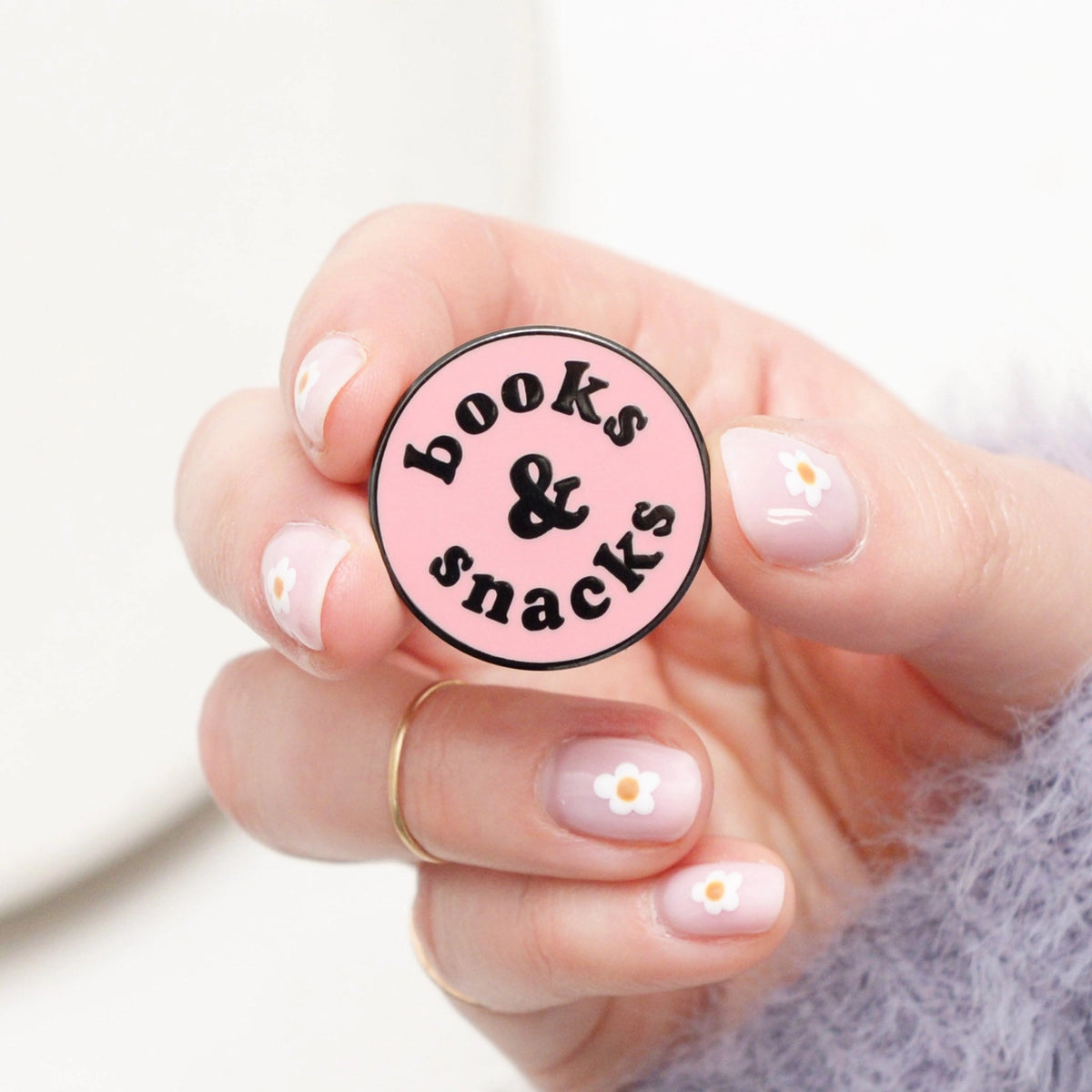 Hand holding a round pink badge with 'books & snacks' text on a light background