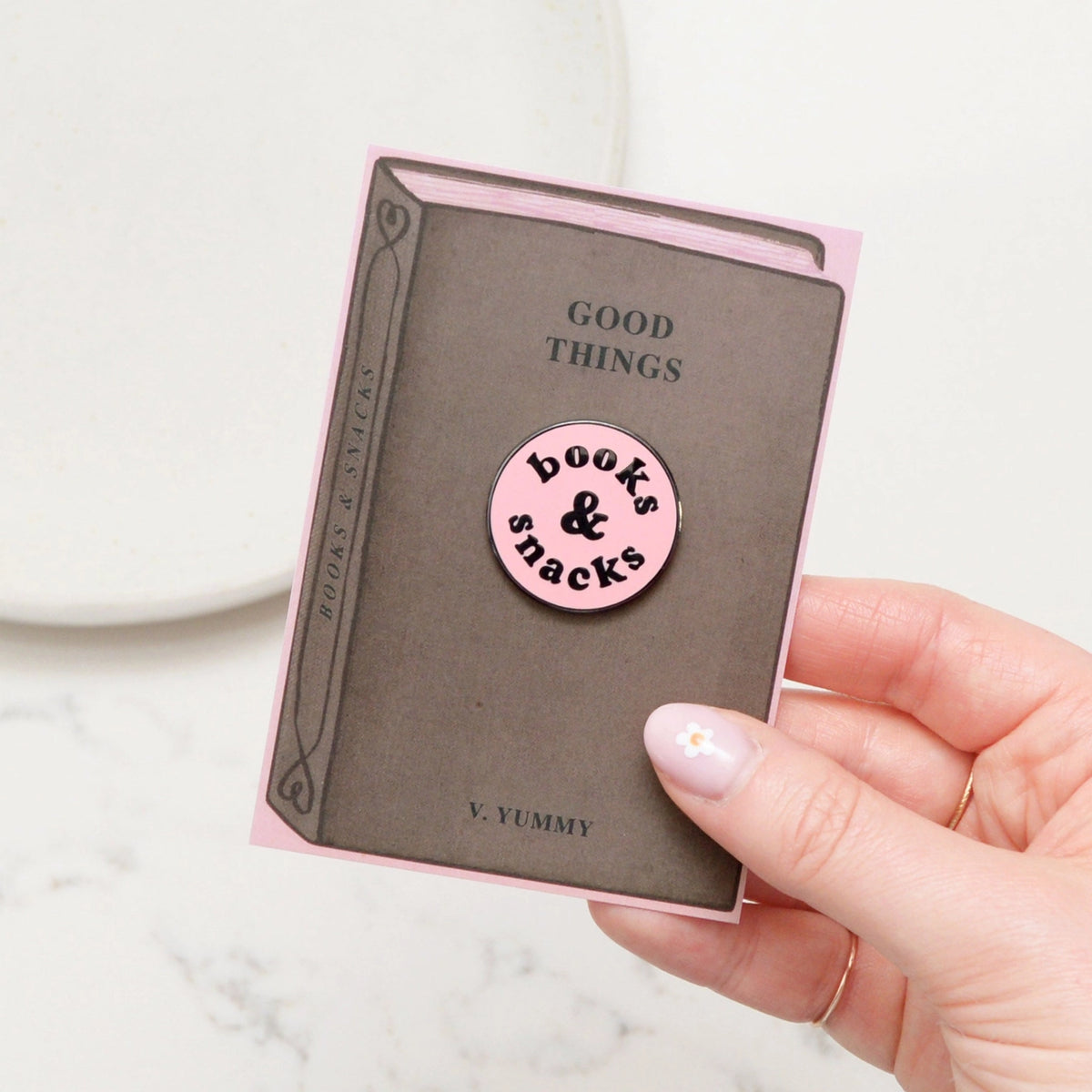 Pink 'books & snacks' enamel pin held over a book with 'Good Things Yummy' text.