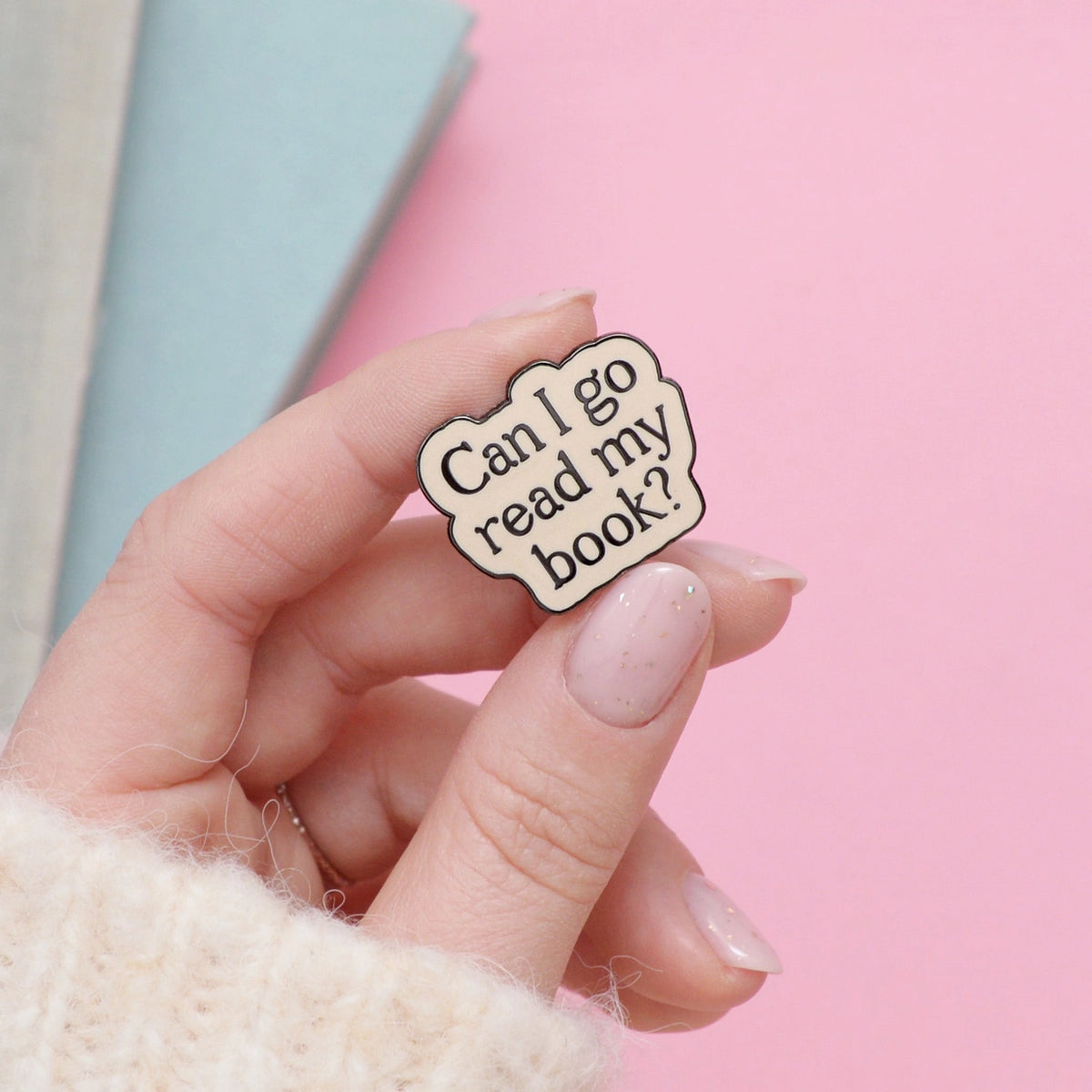 Hand holding a pin with 'Can I go read my book?' text against a pink background