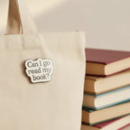 Enamel pin with text 'Can I go read my book?' on a pink and white background