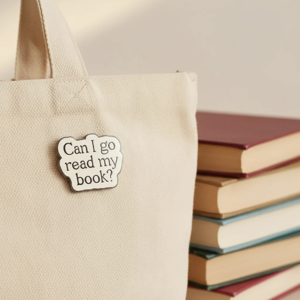 Enamel pin with text 'Can I go read my book?' on a pink and white background