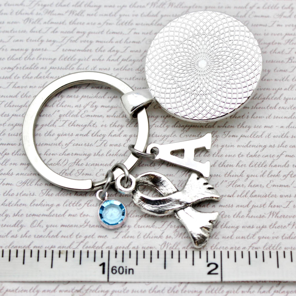 Silver keychain with 'A' and scarf charm on a textured surface with ruler for scale