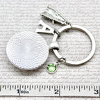 Silver keychain with a round pendant and green gemstone on a textured surface with a ruler for scale.
